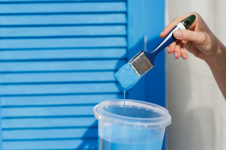 A woman holds a paintbrush dipped in vibrant blue paint, preparing to paint a window, showcasing a creative home improvement project.